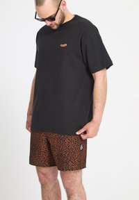 Homme portant un t-shirt noir Volcom et un short orange à motif léopard, regardant vers le bas, avec des cheveux courts et des lunettes de soleil.