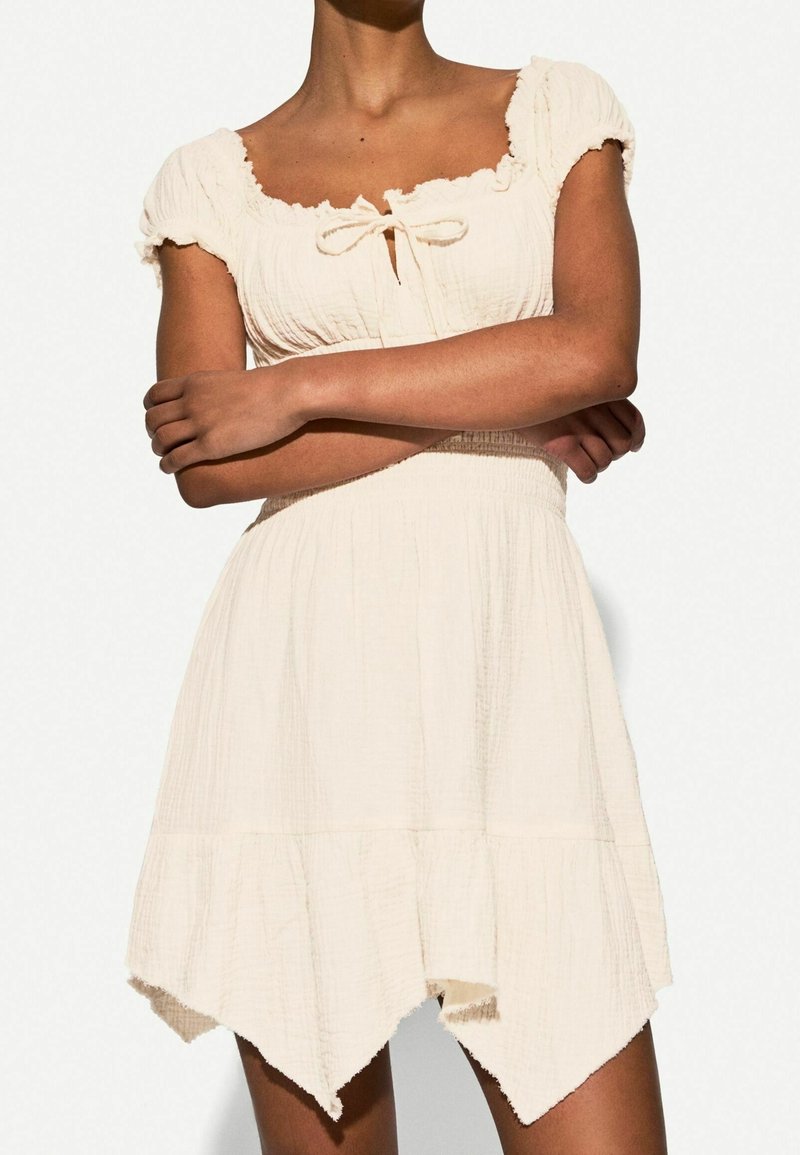 Hellbeiges Kleid aus strukturiertem Stoff. Verfügt über einen quadratischen Ausschnitt, kurze Puffärmel und einen gestuften, asymmetrischen Saum. Bindebanddetails vorne.