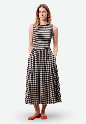 Femme portant une robe midi sans manches à carreaux noirs et blancs avec des poches et des chaussures plates rouges, debout devant un fond uni.