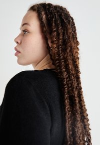 Jersey negro texturizado, perfil lateral de una persona con cabello largo y rizado de color castaño. Las características notables incluyen un piercing en el septo y un pendiente de aro.