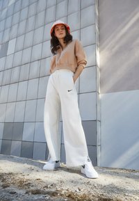 Beige avskåret sweatshirt, hvite vide bukser med Nike-logo, og hvite sneakers på en person mot en moderne geometrisk vegg.