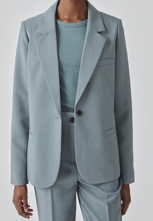 Personne portant une veste de costume bleu clair sur mesure et un pantalon assorti par-dessus un haut bleu côtelé à col rond, sur un fond uni.