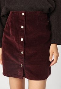 Bordeaux corduroy rok met een knoopsluiting aan de voorkant, voorzien van zilveren metalen knopen, gestructureerde ribbels en een getailleerde vorm.