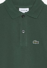 Grön polo-tröja i texturerat tyg, med en tvåknapps öppning och en broderad krokodil-logotyp på vänster bröst.