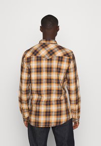 Uomo in piedi con la schiena visibile, che indossa una camicia a quadri arancione, blu navy e bianca a maniche lunghe e pantaloni scuri su uno sfondo semplice.