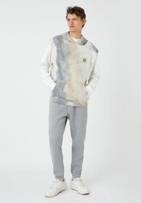 Capuchon, tie-dye sweatshirt in crème- en grijstinten met een buidelzak, gecombineerd met lichtgrijze joggingbroeken en witte sneakers.