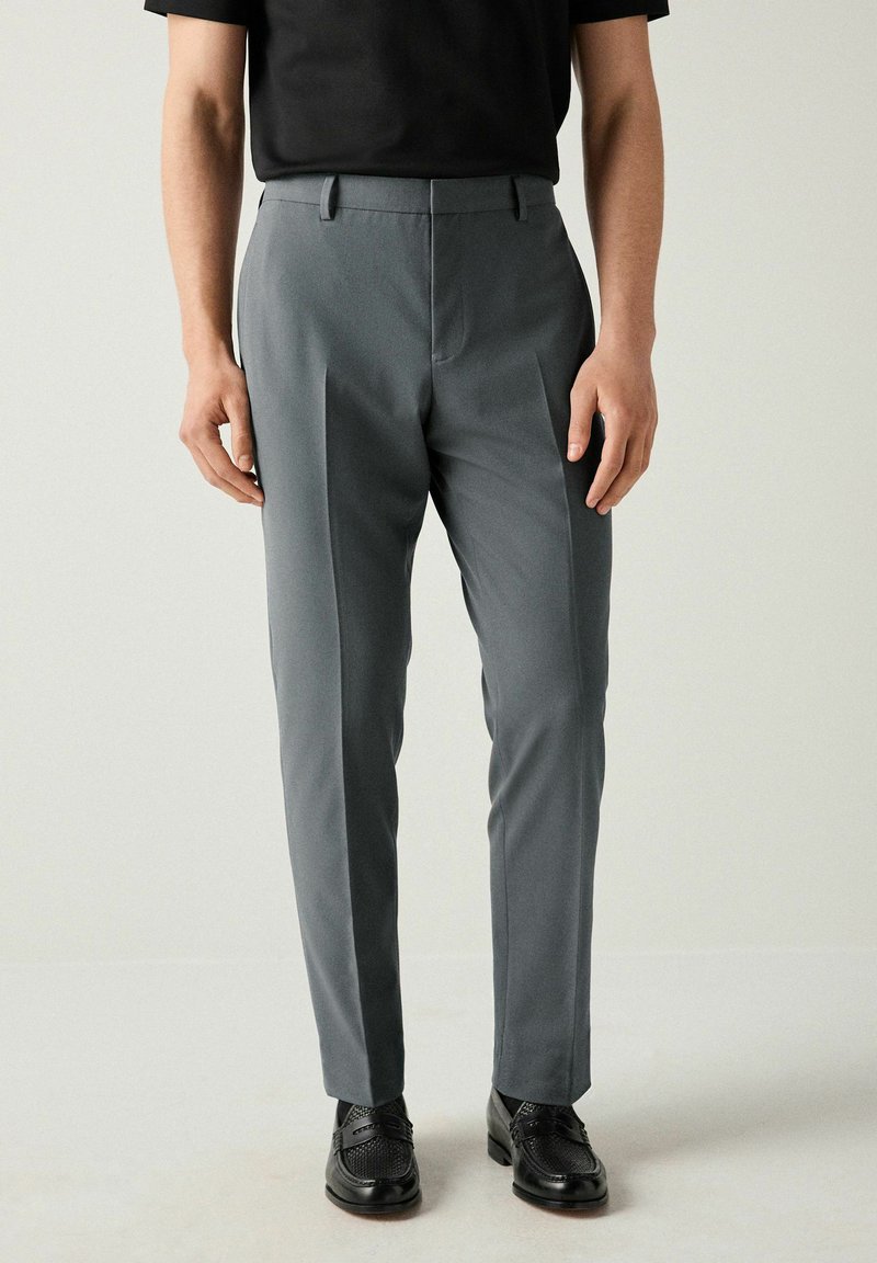 Pantalones gris ajustados hechos de una tela suave, con un corte entallado y pliegues delanteros, combinados con zapatos de vestir negros.