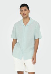 Camisa de manga corta azul claro con cuello, hecha de una tela suave y texturizada. Presenta un cierre de botones en la parte delantera y un corte relajado.