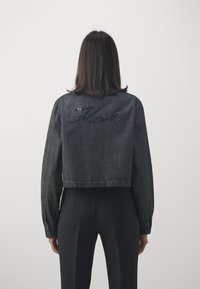 Giacca di jeans nera corta con maniche lunghe, caratterizzata da un logo in paillettes lucide "Karl" sul retro. Texture liscia, design minimalista.
