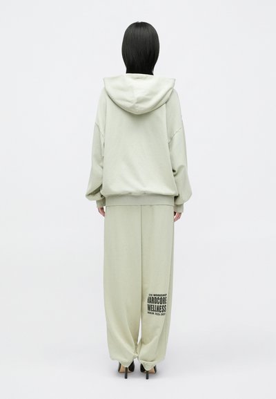 Világos zöld kapucnis pulóver, laza fazonnal és elülső zsebbel; széles szárú nadrággal párosítva, amelynek lábán nyomott logó található.