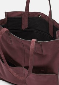 Bordowa zamszowa torba typu tote z czarnym wnętrzem, wyposażona w dwie zewnętrzne kieszenie i dwa paski. Miękka tekstura i prosty design.