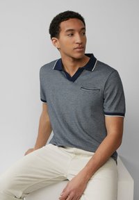 Polo majica s kratkimi rokavi v heather modri barvi z kontrastnim modrim ovratnikom in belimi robovi; vključuje majhen žep na prsih. Model nosi svetle hlače.
