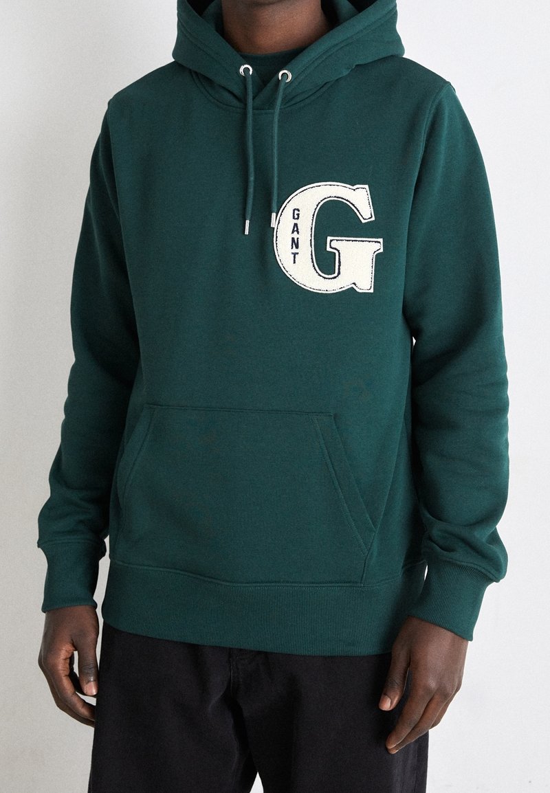 Sweat à capuche vert foncé avec poche avant et grand écusson blanc « G » marqué « GANT » sur la poitrine, porté par une personne à la peau foncée et un pantalon noir.