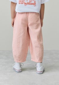 Randiga, vida rosa och vita byxor med sammetsanklar, kombinerade med vita Converse sneakers på en ljusgrå yta.