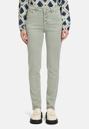 Pantalon slim vert clair avec quatre boutons décoratifs sur le devant, associé à des mocassins crème à glands et une chemise à manches longues à motifs.