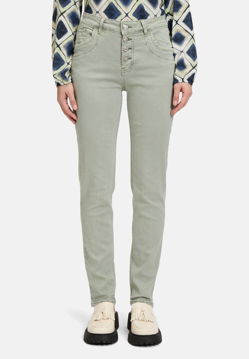 Pantalon slim vert clair avec quatre boutons décoratifs sur le devant, associé à des mocassins crème à glands et une chemise à manches longues à motifs.