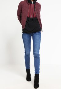 Giacca bomber bordeaux trapuntata sopra una felpa nera con cappuccio, abbinata a jeans skinny blu e stivaletti neri.