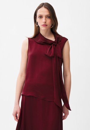 Jeune femme aux cheveux bruns lisses portant un chemisier sans manches bordeaux avec un grand nœud noué au col et une jupe assortie.