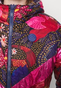 Veste à motif floral coloré avec une fermeture éclair bleu marine et un patch circulaire bleu arborant le logo "Farm Rio Dress in Happiness" sur fond noir.