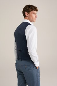 Chaleco navy de textura suave, combinado con una camisa blanca. El chaleco tiene una hebilla en la parte trasera y se lleva con unos pantalones de cuadro azul claro.