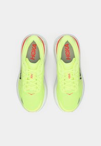 Ζεύγος από νεον κίτρινα παπούτσια τρεξίματος HOKA με λευκές σόλες, κόκκινο λογότυπο στο εσωτερικό και κορδόνια, που φαίνονται από πάνω σε λευκό φόντο.