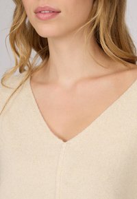 Pull en tricot beige avec un design en V, présentant une texture douce et un subtil éclat, mettant en avant des détails minimalistes et des lignes épurées.