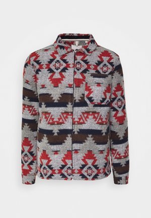 Veste à manches longues avec fermeture éclair et col pointu, présentant un motif tribal géométrique rouge, gris, marine et marron, avec une poche poitrine sur le côté gauche.