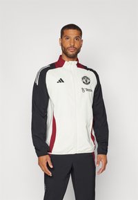 Witte en zwarte trainingsjack met rode accenten en rits, voorzien van het Adidas-logo en het embleem van Manchester United op de borst.