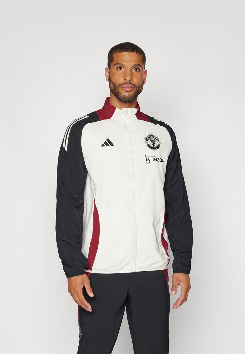 Witte en zwarte trainingsjack met rode accenten en rits, voorzien van het Adidas-logo en het embleem van Manchester United op de borst.
