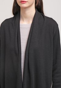 Cardigan nero leggero con un design a drappeggio e apertura frontale. Indossato sopra una maglietta a maniche lunghe grigia chiara a righe. Texture liscia.