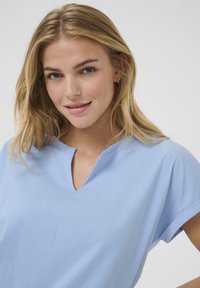 Tricou din bumbac de culoare albastru deschis, cu decolteu în V și mâneci scurte răsucite. Materialul pare moale, cu o textură netedă și o croială lejeră.