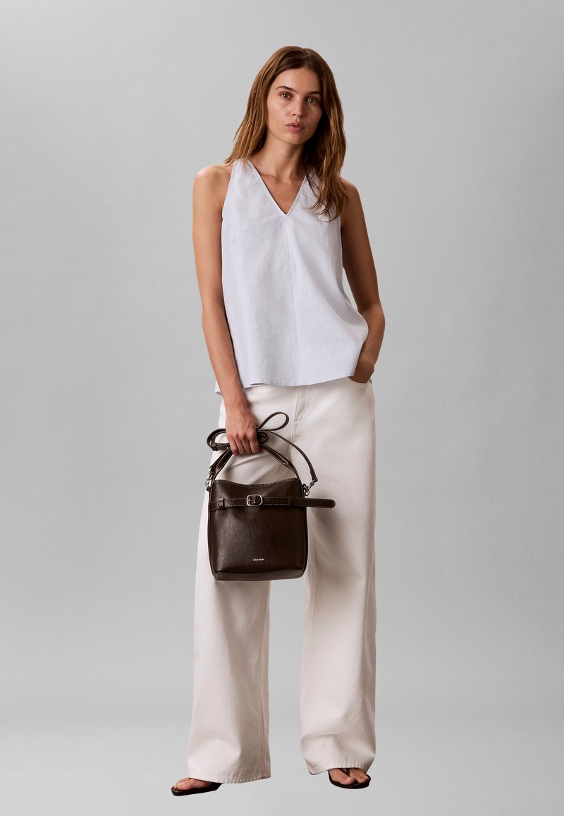 Femme portant un haut blanc sans manches en V et un pantalon large crème, tenant un sac à main en cuir marron foncé avec une boucle, debout sur un fond gris.
