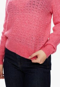 Maglione rosa lavorato a maglia con un motivo testurizzato e dettagli traforati, maniche a costine e orlo svasato, abbinato a jeans blu scuro.