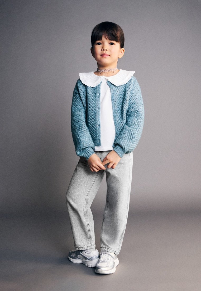 Enfant portant un cardigan en tricot bleu, une chemise blanche à col en dentelle, un pantalon gris et des baskets argentées, debout devant un fond gris.
