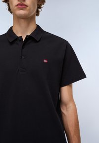 Svart kortermet polo skjorte laget av strukturert stoff, med knappestolpe og et lite brodert emblem i rødt og hvitt på brystet.