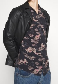 Person, die eine schwarze Lederjacke über einem schwarzen Hemd mit beigen und rosa Drachendesigns sowie blauen Jeans trägt.