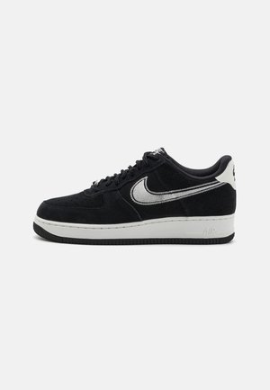 Zapatilla de ante negra con una entresuela blanca, que presenta un logo de Nike en plata y una suela de goma negra. Diseño de cordones con puntera perforada.