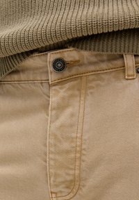 Pantalones de algodón beige con corte recto, con un botón oscuro y costuras visibles. Un suéter texturizado en verde oliva se ve parcialmente arriba.