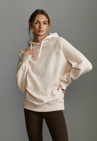 Haut à capuche beige clair avec un cordon de serrage, des manches raglan et une coupe décontractée, associé à des leggings marron foncé. Texture douce et design minimal.
