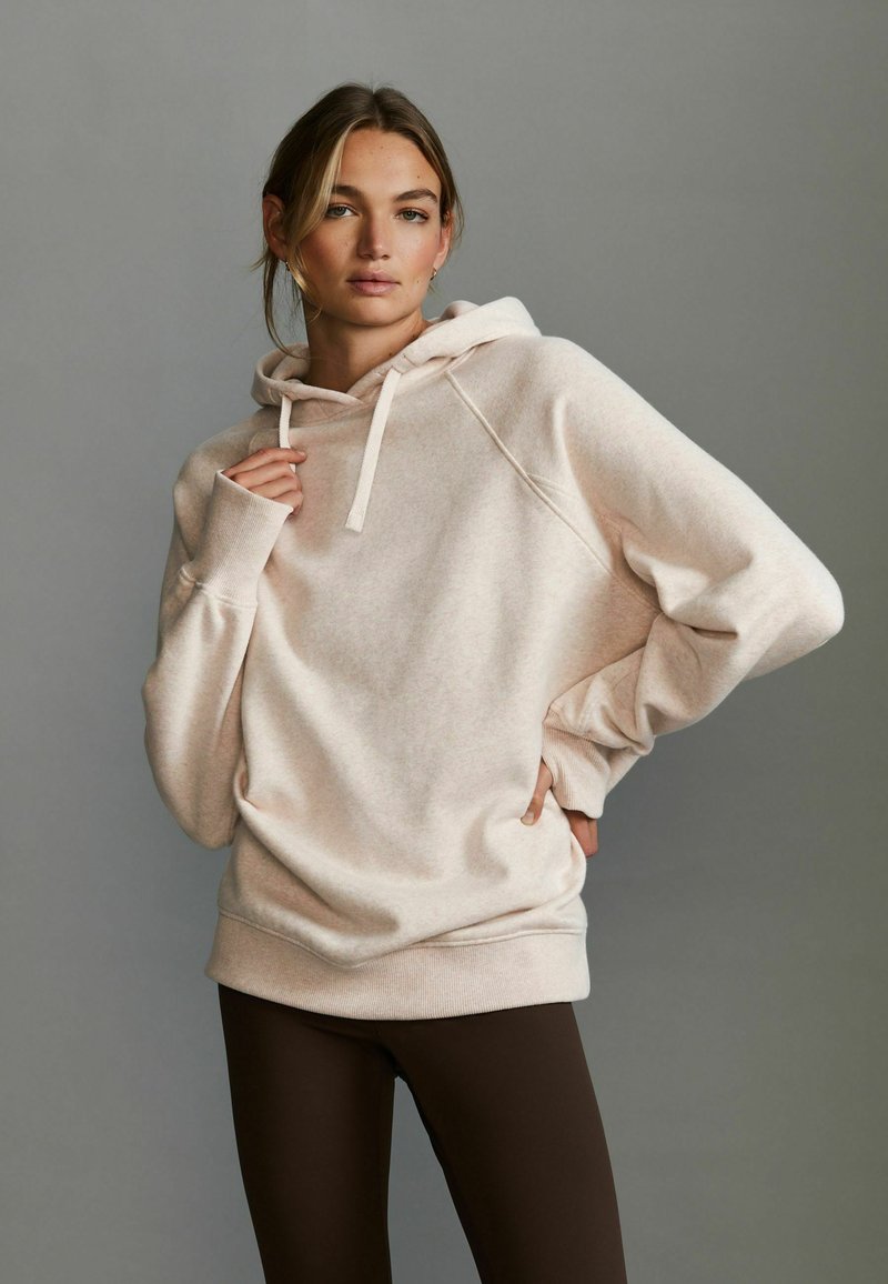 Haut à capuche beige clair avec un cordon de serrage, des manches raglan et une coupe décontractée, associé à des leggings marron foncé. Texture douce et design minimal.