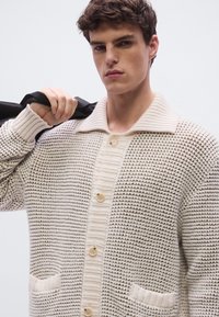 Πλεκτή ζακέτα σε εκρού χρώμα με υφή, διαθέτει ριμπ γιακά, μεγάλα κουμπιά και δύο μπροστινές τσέπες.