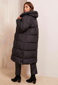 Cappotto lungo imbottito nero con cappuccio, dotato di sezioni trapuntate e chiusure a pressione nella parte inferiore. Indossato sopra pantaloni neri aderenti e stivali.