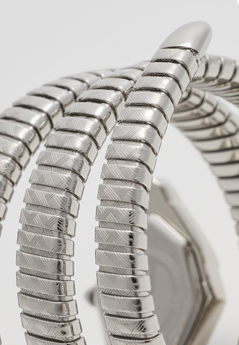 Montre en métal argenté avec un bracelet flexible comportant des segments carrés liés. Le cadran est octogonal avec de subtils motifs gravés.