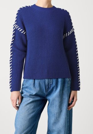 Kvinde iført en kongeblå striktrøje med hvide stingdetaljer langs ærmerne og lyseblå denimjeans.