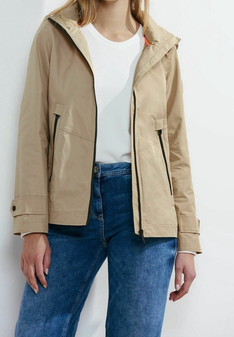 Beige lättviktig jacka med huva, dragkedja fram, två sidofickor och svarta dragkedjedetaljer. Bärs över en vit topp och blå jeans.