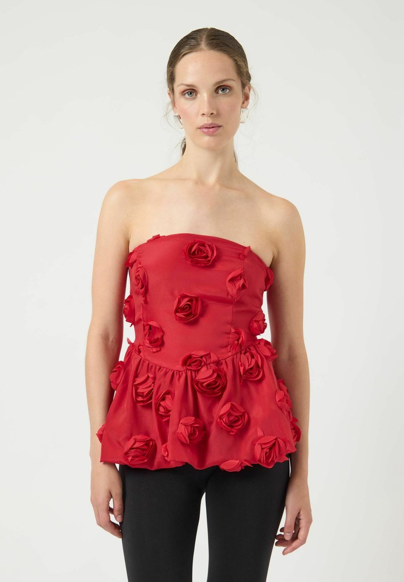 Rød stropløs top med en flagrende kant prydet med stofroser, glat tekstur og en tætsiddende liv. Bæres med sorte bukser.