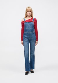 Lichtblauwe denim tuinbroek met verstelbare banden, voorzakken en brede pijpen, gecombineerd met een rood shirt met lange mouwen. Platte zwarte schoenen.