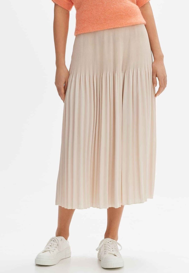 Beige plissé midi rok met een brede, gathered tailleband, gecombineerd met witte sneakers. Zachte textuur, lichte stof valt elegant.