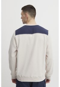 Sudadera de color beige claro con un panel trasero en la parte superior de color azul marino, mangas largas, puños de canalé y un acabado suave y texturizado.