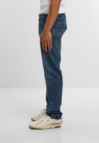 Donkerblauwe denim jeans met een rechte snit, vijf zakken en stiksels. Gecombineerd met lichtbeige sneakers. Zijkant profielweergave.